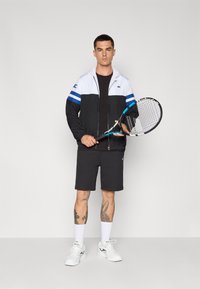 Svart og hvit zip-up jakke med blå striper, svart skjorte, mørke shorts, hvite sko; holder en tenniskølle, viser ben-tatoveringer.