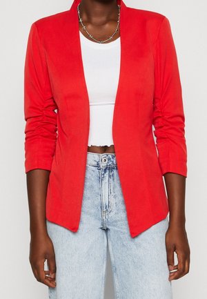 Donkere huidkleurige persoon met een felrode blazer met gerimpelde mouwen, een wit cropped topje, lichtblauwe hoge taille jeans en een zilveren ketting.