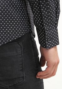 Camicia nera con motivo a fantasia bianca, caratterizzata da maniche lunghe, polsini con bottone e un tessuto testurizzato. Abbinata a jeans scuri.