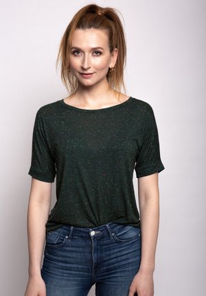 Dunkelgrünes T-Shirt mit kurzen Ärmeln, locker geschnitten, mit mehrfarbigen Sprenkeln und einem runden Ausschnitt. Kombiniert mit blauen Jeans.