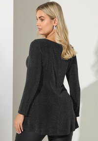 Czarna bluzka z długim rękawem o teksturze z połyskiem, z szerokim dekoltem i rozkloszowanym dołem, w zestawieniu z czarnymi dopasowanymi spodniami.