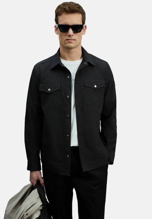 Mann trägt ein schwarzes, geknöpftes Hemd über einem weißen T-Shirt, schwarze Hose und Sonnenbrille, hält eine gefaltete beige Jacke in der linken Hand.