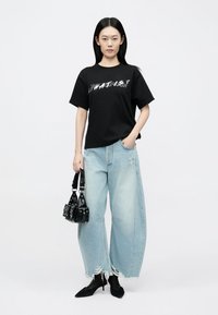 T-shirt gráfica preta com texto branco, combinada com jeans azul claro de perna larga, com bainhas desfiadas. Modelo segura uma pequena mala preta.