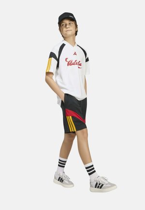 Niño caminando con las manos en los bolsillos, vestido con una camiseta blanca de Adidas con rayas amarillas, pantalones cortos negros con detalles rojos y amarillos, gorra negra y zapatillas.