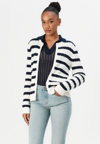 Stripete strikkegenser i hvit og marineblå, med ribbet tekstur, knappelukking og rund hals, kombinert med lyseblå jeans.