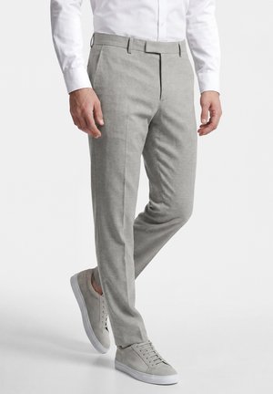 Man draagt lichtgrijze pantalon, wit overhemd en bijpassende grijze sneakers, staat met één voet iets opgetild.