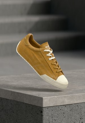 Brown suede sneaker with white rubber toe cap and sole, white and brown laces, positioned above a concrete ledge with stairs in the background.