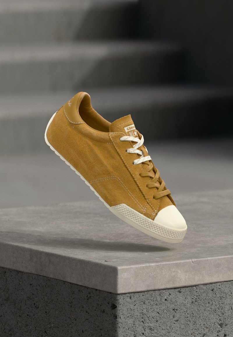 Καφέ σουέντ αθλητικό παπούτσι με λευκή λαστιχένια μύτη και σόλα, λευκά και καφέ κορδόνια, τοποθετημένο πάνω από μια κονκρετική άκρη με σκάλες στο φόντο.