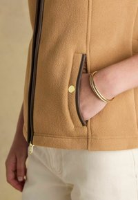Tans Fleece-Jacke mit braunem Reißverschluss und Taschenbesatz. Besitzt einen goldenen Druckknopf an der Tasche und eine glatte Textur. Kombiniert mit cremefarbener Hose.