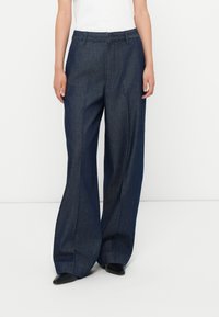 Nő széles szárú sötét farmernadr�ágban, elöl varrással, fekete hegyes orrú cipővel és fehér felsővel áll egy fehér háttér előtt.