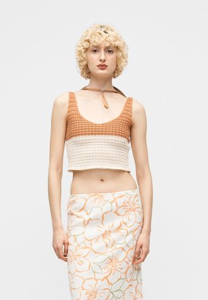 Blond krøllet kvinde iført en ærmeløs brun og beige hæklet croptop med choker-halskæde og et hvidt skørt med orange blomsterprint.