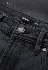 Jeans in denim nero con chiusura a bottone, cuciture visibili e un'etichetta con la scritta "ombre". Presentano una consistenza liscia e un tessuto resistente.