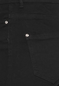 Zwarte denim stof met een achterzak en een zilveren klinknagel detail. De stof is soepel met contrasterende stiksels.