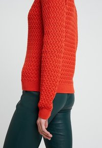 Pull rouge texturé avec un motif en losange, bord-côtes aux poignets et à l'ourlet. Assorti à un pantalon vert ajusté.