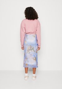 Donna con capelli ricci che indossa un pullover rosa, una gonna midi blu con stampa di angeli, calze bianche e scarpe da ginnastica beige, in piedi di fronte a un muro bianco.