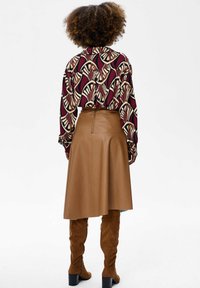 Person with curly hair wearing a patterned long-sleeve shirt, knee-length tan leather skirt, and brown suede knee-high boots, facing away.