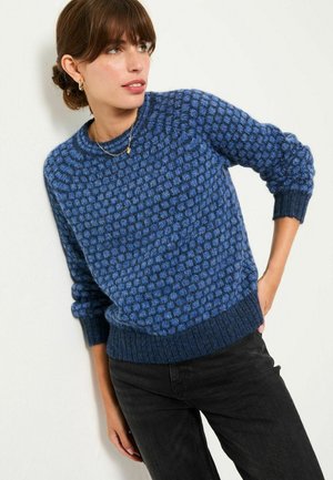 Blauer Strickpullover mit einem strukturierten Bobbelmuster, rundem Halsausschnitt und gerippten Bündchen und Saum. Kombiniert mit schwarzen Hosen.