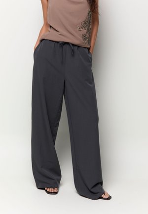 Femme portant un pantalon ample gris foncé à jambes larges avec une taille à cordon et des sandales noires ouvertes à talons carrés.