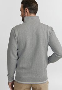 Grauer Zip-Pullover aus weichem Material, mit langen Ärmeln, gerippten Bündchen und einem kontrastierenden gestreiften Saum. Glatte Textur, minimales Design.