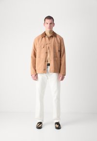 Tan jasje met een kraag, knopen aan de voorkant en twee borstzakken. Draag het over een beige coltrui, gecombineerd met witte broek en zwarte schoenen.