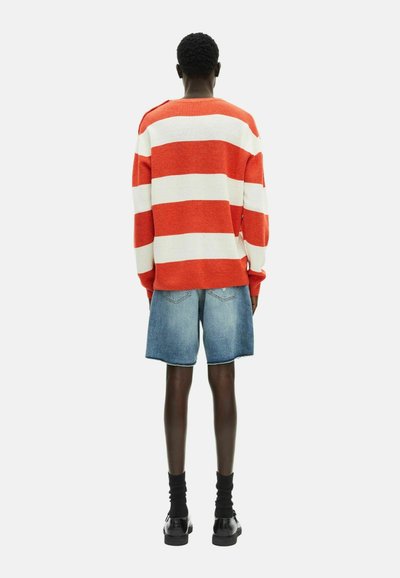 Gestreifter roter und weißer Strickpullover mit fallenden Schultern, kombiniert mit knielangen blauen Denim-Shorts und schwarzen Schuhen, von hinten betrachtet.
