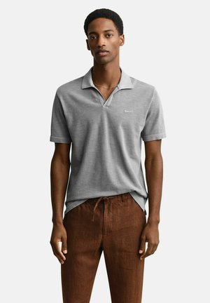 Mann trägt ein hellgraues kurzärmeliges Poloshirt und braune Hose mit Kordelzug, steht vor einem einfarbigen Hintergrund.