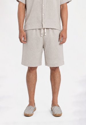Uomo che indossa pantaloncini di lino beige chiaro con coulisse e camicia abbottonata a maniche corte abbinata, abbinata a scarpe slip-on a righe.