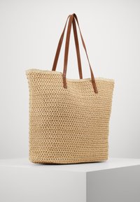 Vævet stråtaske i en naturlig beige farve med brune kunstlæderremme, der har et åbent vævemønster og en rektangulær form.