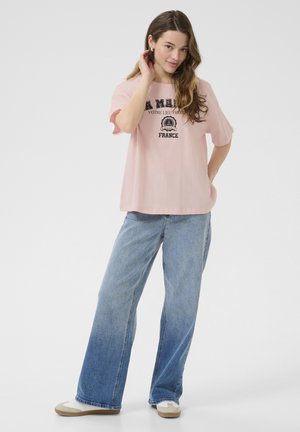 T-shirt en coton rose clair avec un col rond et des manches courtes, affichant un imprimé graphique noir portant les mots "A MIE" et "FRANCE". Porté avec un jean bleu ample.