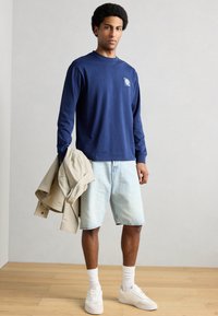 Μπλε ναυτικό πουκάμισο με μακριά μανίκια και γραφικό φοίνικα, ανοιχτό μπλε τζιν σορτς, μπεζ μπουφάν περασμένο στον βραχίονα, λευκά αθλητικά παπούτσια και κάλτσες.