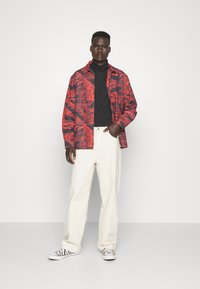 Camicia a righe rossa e nera sovrapposta a un dolcevita nero, abbinata a pantaloni bianchi e scarpe a fantasia. Sfondo minimalista.