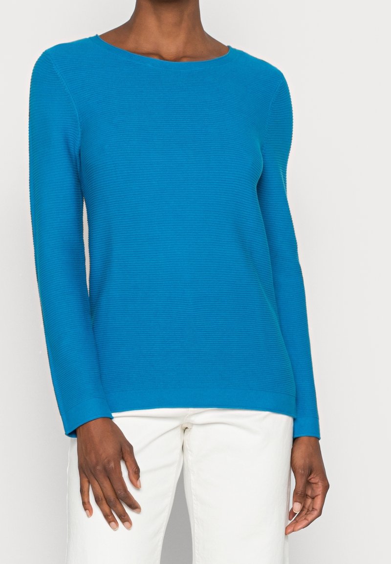 Pull bleu côtelé à manches longues avec un col rond, une texture lisse et des manches ajustées, associé à un pantalon de couleur claire.