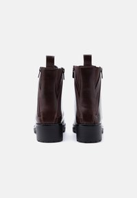 Paire de bottines en cuir marron foncé avec fermeture éclair à l'arrière, languettes de tirage et talons épais noirs, vues de dos sur un fond blanc.