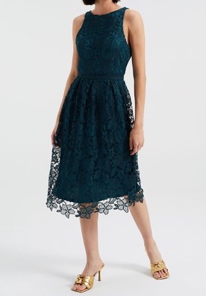 Vestido midi sin mangas de encaje azul oscuro con patrones florales, combinado con mules dorados de tacón alto y punta abierta, en una persona de pie.