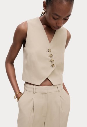 Beige ärmellose Weste mit einem V-Ausschnitt, ausgestattet mit vier dekorativen Knöpfen und einem cropped Schnitt, kombiniert mit passenden hochgeschnittenen Hosen.
