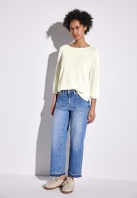 Maglione giallo chiaro, dalla consistenza morbida, con maniche a tre quarti, abbinato a jeans in denim a gamba larga di colore blu chiaro e sneakers beige.