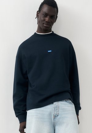 Άνδρας φορώντας σκούρο μπλε φούτερ με μικρό μπλε λογότυπο "HUGO" και ανοιχτά μπλε jeans, στέκεται μπροστά σε απλό ανοιχτό φόντο.