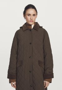 Brun quiltet frakke med krave og sorte knapper, prydet med brune corduroy-detaljer på lommer og manchetter; tekstureret stof.