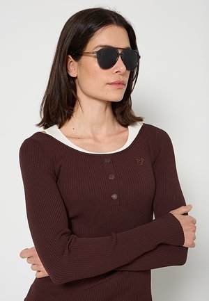 Donna con capelli corti e scuri che indossa occhiali da sole neri e una maglia aderente a coste marrone scuro a maniche lunghe con bottoni e una canottiera bianca.