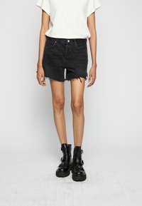 Pantalones cortos de mezclilla negros con un dobladillo deshilachado, combinados con botas negras hasta el tobillo que tienen correas y hebillas. Una camiseta blanca de gran tamaño completa el conjunto.