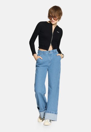 Osoba ubrana w oversize'owe jasnoniebieskie jeansy z mankietami, czarny krótki sweter na zamek, okulary przeciwsłoneczne oraz białe trampki z czarnymi paskami.