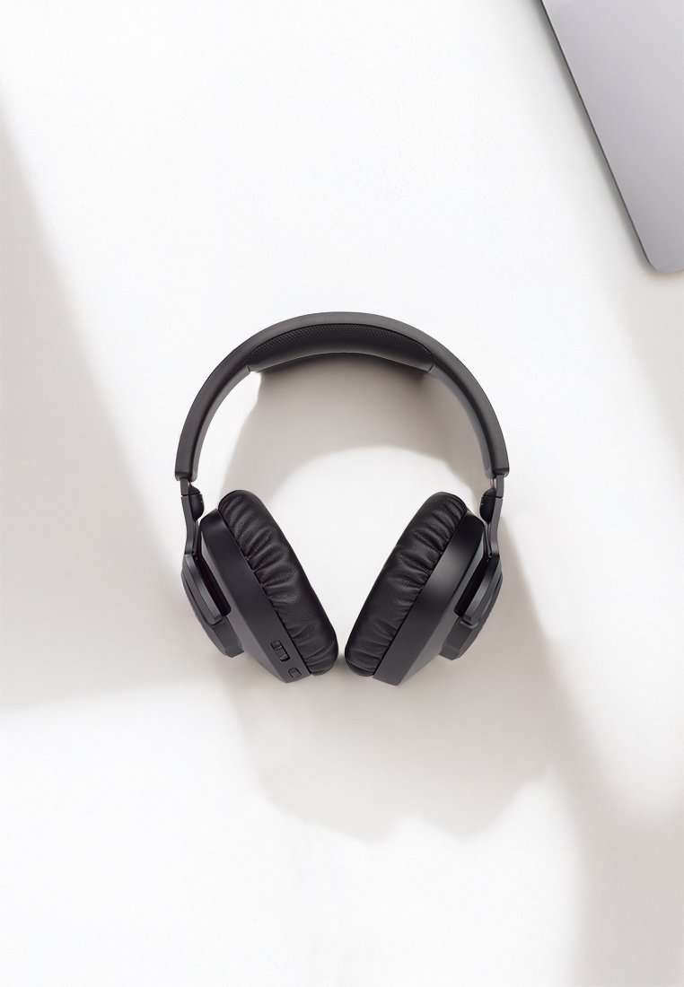 Cuffie wireless over-ear nere appoggiate su una scrivania bianca vicino all'angolo di un laptop argento, con ombre morbide visibili.