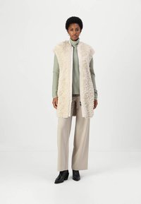 Gilet lungo in pelliccia sintetica color crema, con un'esterno testurizzato, apertura frontale e foderato leggermente beige, indossato sopra un dolcevita verde e pantaloni beige.