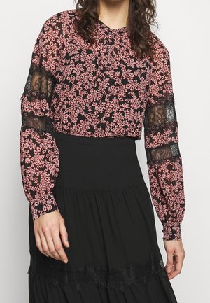 Kvinde iført en langærmet rosa og sort blomstret bluse med sorte blondeindsatser på ærmerne og et sort nederdel med høj talje.