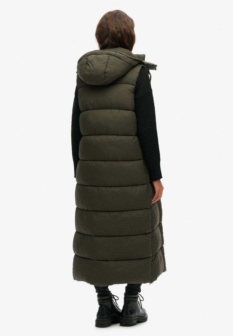 Langer, olivgrüner Puffer-Weste mit Kapuze, die breite horizontale Abschnitte und einen leicht strukturierten Stoff aufweist. Über einem schwarzen Oberteil getragen.