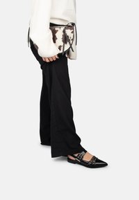 Persona che indossa pantaloni neri a gamba larga, maglione bianco, ballerine nere con punte appuntite e borchie, e tiene una borsa a tracolla in pelle di mucca marrone e bianca.