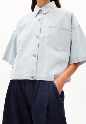 Světlomodrá košile s krátkými rukávy na knoflíky s kapsou na hrudi, nošená s tmavě modrými vysoce pasovými kalhotami s plisováním, ruka v kapse.