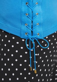 Blå topp med snörningsdesign som har guldfärgade öglor, tillsammans med svarta byxor täckta av vita prickar. Texturerad tyg synlig.