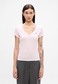 Ροζ ανοιχτό V-neck μπλούζα με κοντό μανίκι, φτιαγμένη από μαλακό ύφασμα. Μικρή λεπτομέρεια λογότυπου στο αριστερό στήθος. Συνδυάζεται με γκρι παντελόνι με ρίγες.