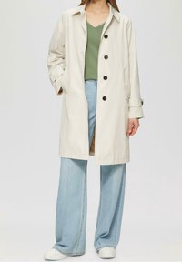 Jasnobeżowy trencz z klasycznym kołnierzem, pięcioma ciemnymi guzikami i dwoma bocznymi kieszeniami, zestawiony z zielonym topem i szerokimi niebieskimi dżinsami.